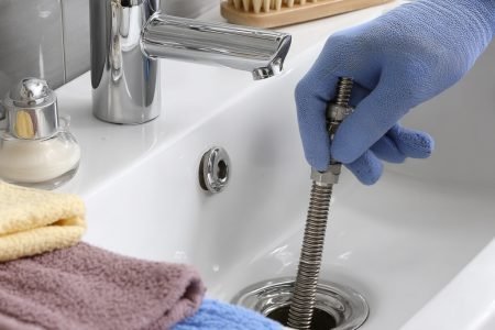 A bathroom sink with drain auger equipment inserted. A technician’s gloved hand is turning the auger handle. Plumber tools and cleaning cloths are placed beside the sink. --v 7 Job ID: 38027e15-b159-4116-829f-5bce786b26e6