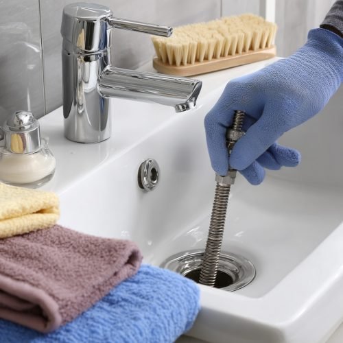 A bathroom sink with drain auger equipment inserted. A technician’s gloved hand is turning the auger handle. Plumber tools and cleaning cloths are placed beside the sink. --v 7 Job ID: 38027e15-b159-4116-829f-5bce786b26e6