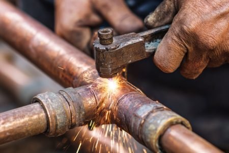 Close-up of copper pipes being replaced next to old galvanized pipes. A technician’s hands hold a pipe wrench, tightening the new fittings. Sparks of light from a small torch in the background. --v 7 Job ID: f733693a-7d56-4790-a9ba-738e9422b99b